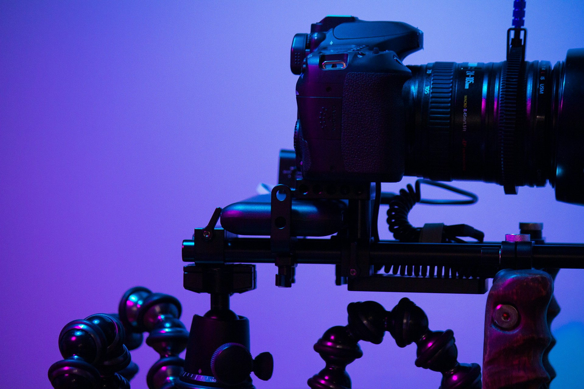 A camera mounted on a tripod setup, featuring a complex rig with multiple adjustable joints. The lighting is a blend of purple and blue hues, casting a futuristic and tech-savvy ambiance.