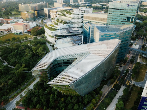 An aerial view of a modern urban area featuring several large, uniquely designed buildings surrounded by lush greenery. The central building has a distinctive, angular shape with a roof partially covered in solar panels. Additional structures with curved designs and glass facades are visible in the vicinity, forming a cohesive architectural setting.