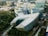 An aerial view of a modern urban area featuring several large, uniquely designed buildings surrounded by lush greenery. The central building has a distinctive, angular shape with a roof partially covered in solar panels. Additional structures with curved designs and glass facades are visible in the vicinity, forming a cohesive architectural setting.