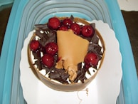 A round dessert topped with glossy red cherries and dark chocolate shavings, adorned with a light brown chocolate curl. It is presented on a white decorative paper within a turquoise container.