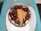 A round dessert topped with glossy red cherries and dark chocolate shavings, adorned with a light brown chocolate curl. It is presented on a white decorative paper within a turquoise container.