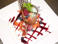 A gourmet dessert featuring a chocolate cylinder filled with various berries including raspberries, blueberries, and strawberries. There is a decorative chocolate lattice structure and fresh mint leaves on top, all presented on a white plate with artistic red sauce drizzle and small dollops of cream.
