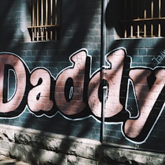 Large graffiti letters spelling 'Daddy' are painted on a brick wall. Shadows and sunlight create a pattern of contrasting light and dark across the letters, and there are barred windows above.
