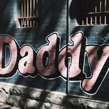 Large graffiti letters spelling 'Daddy' are painted on a brick wall. Shadows and sunlight create a pattern of contrasting light and dark across the letters, and there are barred windows above.