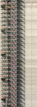 An industrial structure with a scaffolding system against a large concrete wall. The scaffolding features metal beams, platforms, and red sections, casting intricate shadows onto the wall. The concrete surface is smooth with subtle vertical lines.