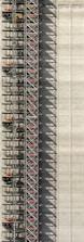 An industrial structure with a scaffolding system against a large concrete wall. The scaffolding features metal beams, platforms, and red sections, casting intricate shadows onto the wall. The concrete surface is smooth with subtle vertical lines.