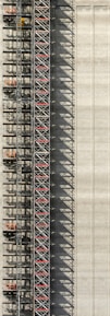 An industrial structure with a scaffolding system against a large concrete wall. The scaffolding features metal beams, platforms, and red sections, casting intricate shadows onto the wall. The concrete surface is smooth with subtle vertical lines.
