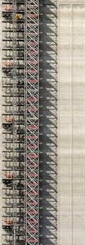 An industrial structure with a scaffolding system against a large concrete wall. The scaffolding features metal beams, platforms, and red sections, casting intricate shadows onto the wall. The concrete surface is smooth with subtle vertical lines.
