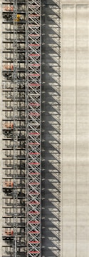 An industrial structure with a scaffolding system against a large concrete wall. The scaffolding features metal beams, platforms, and red sections, casting intricate shadows onto the wall. The concrete surface is smooth with subtle vertical lines.
