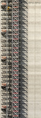An industrial structure with a scaffolding system against a large concrete wall. The scaffolding features metal beams, platforms, and red sections, casting intricate shadows onto the wall. The concrete surface is smooth with subtle vertical lines.