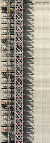 An industrial structure with a scaffolding system against a large concrete wall. The scaffolding features metal beams, platforms, and red sections, casting intricate shadows onto the wall. The concrete surface is smooth with subtle vertical lines.