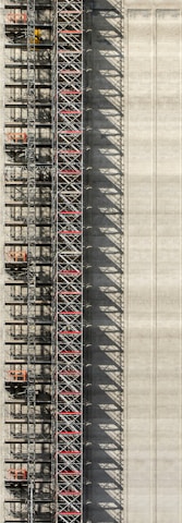 An industrial structure with a scaffolding system against a large concrete wall. The scaffolding features metal beams, platforms, and red sections, casting intricate shadows onto the wall. The concrete surface is smooth with subtle vertical lines.