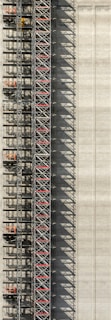 An industrial structure with a scaffolding system against a large concrete wall. The scaffolding features metal beams, platforms, and red sections, casting intricate shadows onto the wall. The concrete surface is smooth with subtle vertical lines.