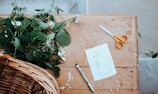 A wooden table features a mixture of creative and natural elements. On the table, there is a pair of orange-handled scissors, a black marker, and a sheet of paper with botanical sketches. Fresh green foliage and small white flowers lie on the table, with a wicker basket partially visible at one corner.