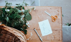 A wooden table features a mixture of creative and natural elements. On the table, there is a pair of orange-handled scissors, a black marker, and a sheet of paper with botanical sketches. Fresh green foliage and small white flowers lie on the table, with a wicker basket partially visible at one corner.