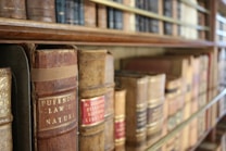Rows of old, leather-bound books with embossed titles sit on a wooden bookshelf. The spines show signs of aging and wear, hinting at the historical significance and the vintage quality of the collection.
