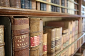 Rows of old, leather-bound books with embossed titles sit on a wooden bookshelf. The spines show signs of aging and wear, hinting at the historical significance and the vintage quality of the collection.