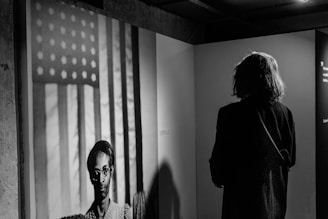 A person stands in an exhibition space, viewing a black and white photograph of an individual in front of a backdrop featuring the American flag. The space around them is dimly lit, focusing attention on both the observer and the photograph.