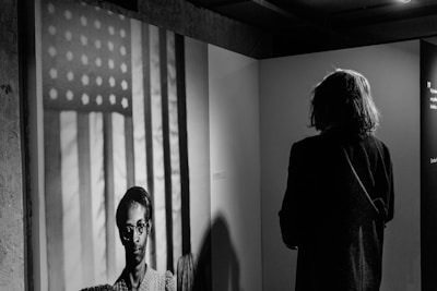 A person stands in an exhibition space, viewing a black and white photograph of an individual in front of a backdrop featuring the American flag. The space around them is dimly lit, focusing attention on both the observer and the photograph.