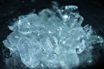 A close-up view of multiple ice cubes clustered together, reflecting light and creating a cool, frosty appearance. The surface of the ice appears smooth and glossy, with variations in transparency adding depth to the composition.