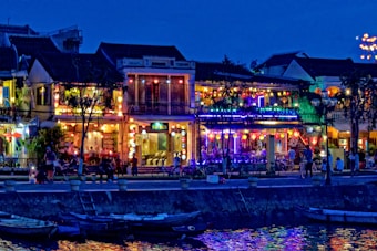 A vibrant nighttime street scene with colorful lanterns adorning the exterior of buildings. The reflections of the lights shimmer in the water of a nearby canal. People are walking along the street, and bicycles are parked near the sidewalk. The atmosphere is festive with an array of illuminated signs and decorations.