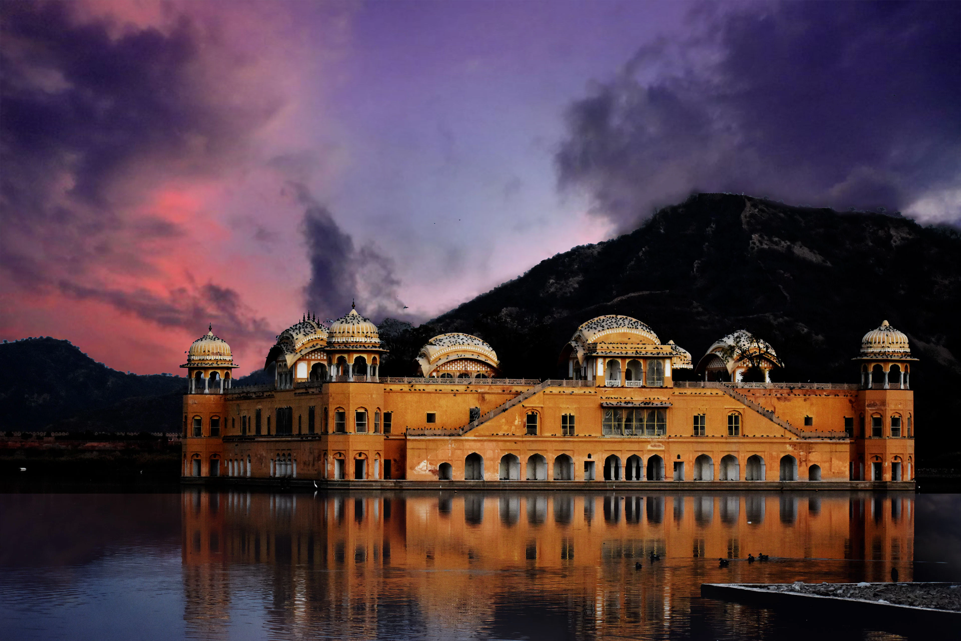 Jal Mahal reflected in the lake under a dramatic violet and pink sunset sky with hills behind.