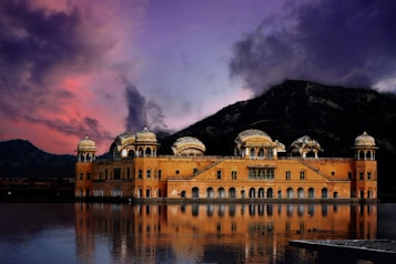 An ornate palace stands majestically on a calm lake, surrounded by picturesque mountains during a vivid sunset. The sky is painted with deep shades of purple and pink, reflecting softly on the water. Architectural features include multiple domes and intricate details.