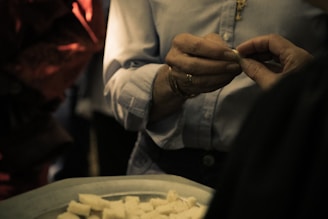 Two hands are engaged in an exchange, with one hand passing a small object to another hand. The person holding the object appears to be wearing a light-colored, button-up shirt with a necklace visible. A tray with small cube-shaped pieces, possibly food, is in the lower part of the image. The lighting is dim, creating a warm, intimate atmosphere.