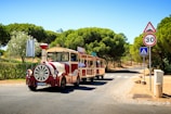 A small tourist train with a vintage appearance travels down a cobblestone path surrounded by lush green trees and shrubs. The train is painted in red and cream colors and has 'Traint' written on it. There are several passengers on board enjoying the ride. Traffic signs, including a speed limit of 30 km/h and a pedestrian crossing sign, are visible along the road.