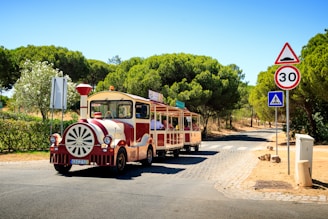 A small tourist train with a vintage appearance travels down a cobblestone path surrounded by lush green trees and shrubs. The train is painted in red and cream colors and has 'Traint' written on it. There are several passengers on board enjoying the ride. Traffic signs, including a speed limit of 30 km/h and a pedestrian crossing sign, are visible along the road.