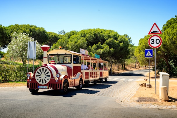 A small tourist train with a vintage appearance travels down a cobblestone path surrounded by lush green trees and shrubs. The train is painted in red and cream colors and has 'Traint' written on it. There are several passengers on board enjoying the ride. Traffic signs, including a speed limit of 30 km/h and a pedestrian crossing sign, are visible along the road.