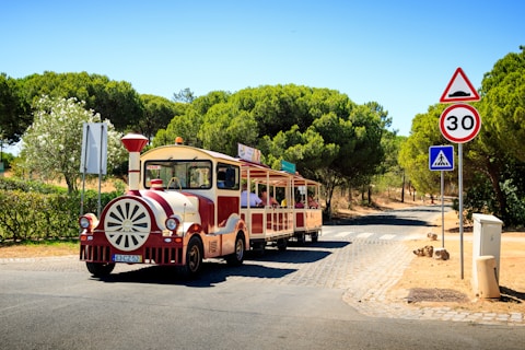 A small tourist train with a vintage appearance travels down a cobblestone path surrounded by lush green trees and shrubs. The train is painted in red and cream colors and has 'Traint' written on it. There are several passengers on board enjoying the ride. Traffic signs, including a speed limit of 30 km/h and a pedestrian crossing sign, are visible along the road.