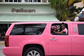 A bright pink limousine SUV is parked on the street in front of a building with a sign that reads 'Pelican.' Two children are visible through the open window of the limo, one wearing a pink headband and the other in a pink and white shirt. Palm tree reflections can be seen on the black tinted windows of the vehicle.