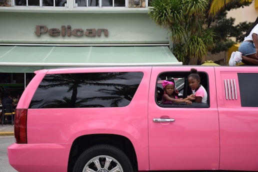 A bright pink limousine SUV is parked on the street in front of a building with a sign that reads 'Pelican.' Two children are visible through the open window of the limo, one wearing a pink headband and the other in a pink and white shirt. Palm tree reflections can be seen on the black tinted windows of the vehicle.