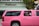 A bright pink limousine SUV is parked on the street in front of a building with a sign that reads 'Pelican.' Two children are visible through the open window of the limo, one wearing a pink headband and the other in a pink and white shirt. Palm tree reflections can be seen on the black tinted windows of the vehicle.