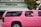 A bright pink limousine SUV is parked on the street in front of a building with a sign that reads 'Pelican.' Two children are visible through the open window of the limo, one wearing a pink headband and the other in a pink and white shirt. Palm tree reflections can be seen on the black tinted windows of the vehicle.