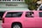 A bright pink limousine SUV is parked on the street in front of a building with a sign that reads 'Pelican.' Two children are visible through the open window of the limo, one wearing a pink headband and the other in a pink and white shirt. Palm tree reflections can be seen on the black tinted windows of the vehicle.