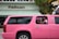 A bright pink limousine SUV is parked on the street in front of a building with a sign that reads 'Pelican.' Two children are visible through the open window of the limo, one wearing a pink headband and the other in a pink and white shirt. Palm tree reflections can be seen on the black tinted windows of the vehicle.