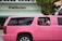 A bright pink limousine SUV is parked on the street in front of a building with a sign that reads 'Pelican.' Two children are visible through the open window of the limo, one wearing a pink headband and the other in a pink and white shirt. Palm tree reflections can be seen on the black tinted windows of the vehicle.