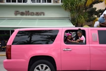 A bright pink limousine SUV is parked on the street in front of a building with a sign that reads 'Pelican.' Two children are visible through the open window of the limo, one wearing a pink headband and the other in a pink and white shirt. Palm tree reflections can be seen on the black tinted windows of the vehicle.
