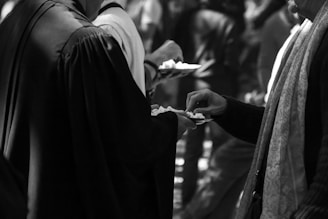 Two individuals are engaged in a gesture of sharing or receiving small items on plates. One person, clothed in dark, flowing garments, holds a plate while another extends their hand towards it. The setting appears to be crowded, suggesting a public event or gathering.
