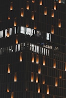 A tall building at night with a grid-like facade is illuminated by warm orange lights. The windows display white light from the interior, revealing some office spaces. The glowing lights create a pattern against the dark exterior.