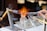 A detailed depiction of a glassblowing process, where a person uses tongs to shape hot glass. Two burners are positioned on either side, with one flame prominently visible. The setup is on a white work surface with various tools in view.