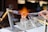 A detailed depiction of a glassblowing process, where a person uses tongs to shape hot glass. Two burners are positioned on either side, with one flame prominently visible. The setup is on a white work surface with various tools in view.