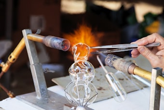 A detailed depiction of a glassblowing process, where a person uses tongs to shape hot glass. Two burners are positioned on either side, with one flame prominently visible. The setup is on a white work surface with various tools in view.