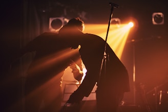 Silhouettes of two people on stage in front of vibrant backlighting, creating dramatic shadows and a warm, atmospheric ambiance. The scene is set with musical equipment, indicating a performance or concert environment.