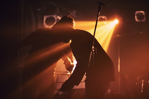 Silhouettes of two people on stage in front of vibrant backlighting, creating dramatic shadows and a warm, atmospheric ambiance. The scene is set with musical equipment, indicating a performance or concert environment.