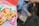 A bag of assorted hard candies with colorful wrappers is prominently displayed in the foreground. The wrappers are predominantly yellow, pink, orange, and green. In the background, there is a blurred image of a person holding an object, possibly a phone, near a campfire.