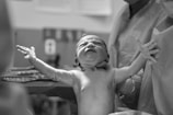 A newborn baby is being held by medical staff in a hospital setting. The baby is extending its arms and appears to have just been delivered. The scene is captured in black and white, highlighting the textures and emotions of the moment.
