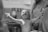 A newborn baby is being held by medical staff in a hospital setting. The baby is extending its arms and appears to have just been delivered. The scene is captured in black and white, highlighting the textures and emotions of the moment.